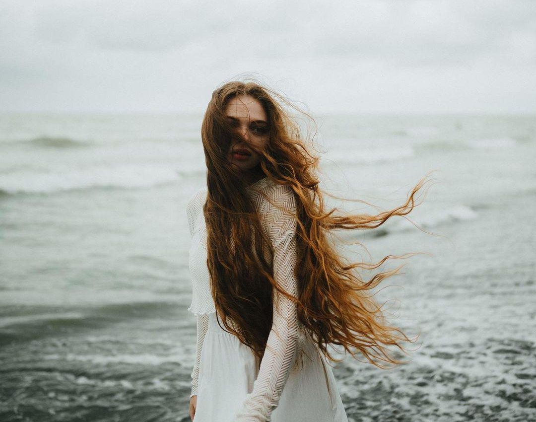 Frau mit langen, windgepeitschten Haaren am Strand – unterstreicht den Einfluss von Umweltfaktoren auf das Haarwachstum und die Haargesundheit.